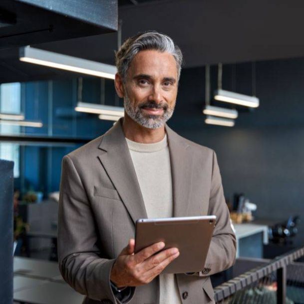 Ein Senior Sales Manager im Anzug steht mit einem Tablet in der Hand in einem Büro